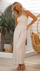 Beige asymmetrical strapless jumpsuit with pleated high waist, front view on a patio.