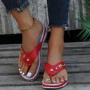 Red wedge thong sandal with perforated upper strap and stud embellishments, cushioned low flatform sole, angled front view on feet.