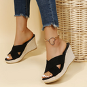Black cross-strap wedge mule sandals with woven espadrille platform, worn with distressed jeans, angled front view.