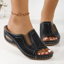 Black open toe wedge mule sandals with white contrast topstitching and elasticated side gusset panel, worn on foot with gold chain anklet, angled front view.