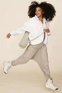 Woman in a beige jumpsuit with large pockets and a white fleece jacket, angled full-body view.