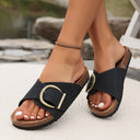 Black flat slip-on sandals with antique-gold adjustable buckle strap and cork sole, angled side view on feet with outdoor background.