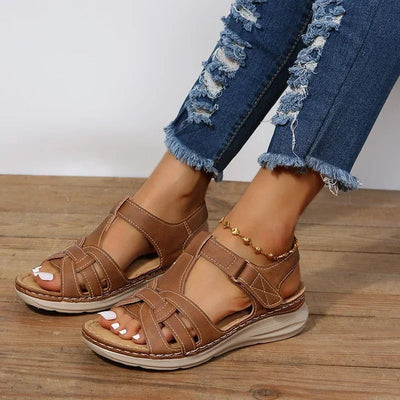 Tan brown low wedge sandals with woven strappy upper, velcro strap closure and cream cushioned sole, worn with distressed denim jeans, angled front view on wooden surface.