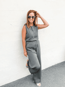 Woman in gray sleeveless wide-leg jumpsuit with sunglasses and necklace, full-body angled view.