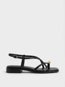 Black strappy flat sandal with crossover toe straps, gold orb accent and ankle buckle, side profile on white background.
