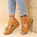 Tan low wedge platform sandals with two adjustable hook-and-loop straps and perforated upper, open-toe, styled on feet with distressed jeans — front view.