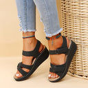 Black low wedge platform sandals with adjustable hook-and-loop straps and contrast stitching, styled with distressed jeans — angled front view.