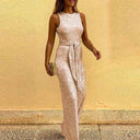 Rose gold sequin sleeveless jumpsuit with waist tie, angled front view against yellow wall.