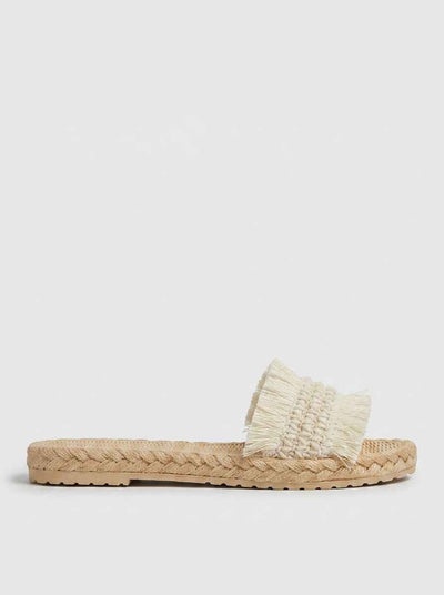 Cream fringed woven strap flat espadrille slide sandal on braided jute sole, side view on white background.
