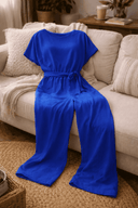 Royal blue wide-leg belted jumpsuit with short sleeves, front view on a light beige couch.