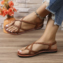 Brown braided flat sandals with woven cross-front straps, slingback ankle strap and cushioned footbed, angled top-side view on wooden surface.