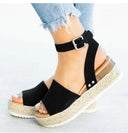 Black espadrille flatform sandal with ankle buckle strap and woven jute platform sole, side view on foot.