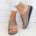 Taupe open-toe wedge mule sandals with wide crosshatch-texture toe-band and dark ridged rubber sole, worn with white trousers, angled front view.