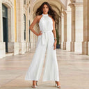 White pleated halter neck jumpsuit with wide legs and tie waist, front view on arched walkway.