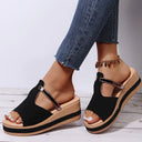 Black platform wedge sandal with broad vamp strap and metallic buckle ankle strap, open-toe silhouette, cushioned tan footbed — front three-quarter view on white wooden floor.