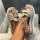 Beige woven platform wedge sandals with large fabric bow and braided jute-style sole, open-toe slip-on, angled side view worn on foot.