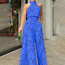 Blue halter-neck jumpsuit with black abstract face print, pockets, and wide legs, angled front view.