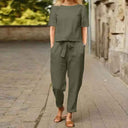 Olive green two-piece jumpsuit with short-sleeve top and tie-waist pants, front view on a brick path.