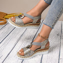 Black and gold open-toe espadrille wedge sandals with ring-hardware ankle strap and gold chain accent, styled on feet with jeans, angled view.