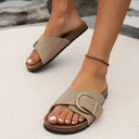 Taupe flat slip-on sandals with wide adjustable buckle strap and cork-look flatbed sole, angled view on feet.
