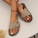 Taupe flat slip-on sandals with wide buckle strap and cork-texture flatbed sole, styled on feet photographed from above.