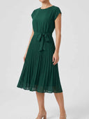 Clara dark green cap sleeve pleated midi dress with tied bow waist, front angled view showing full accordion-pleated A-line skirt — green wedding guest outfit.