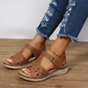 Tan brown low wedge sandals with woven strappy upper, velcro strap closure and cream cushioned sole, worn with distressed denim jeans, angled front view on wooden surface.