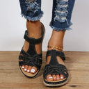 Black low wedge sandals with woven strappy upper, white contrast stitching and cream cushioned sole, angled front view on wooden surface.