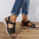 Black low wedge sandals with velcro strap, white contrast stitching and cream cushioned wedge sole, angled front view on wooden floor.