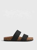 Black two-strap flat slide sandals with cork footbed and rubber outsole, side view on white background.
