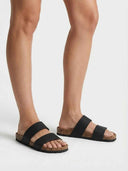 Black two-strap flat slide sandals with cork footbed worn on feet, angled view showing double-strap detail.