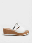 White cushioned wedge mule sandal with wide toe-band and metallic silver cross-strap accent on a tan platform wedge sole, side view.