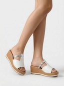 White wedge mule sandals worn on feet showing silver strap detail and tan platform wedge sole, angled view.