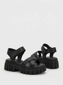 Black chunky platform sandals with crossover toe straps, hook-and-loop ankle strap and thick lug sole platform, angled front view.