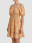 Daisy off-shoulder smocked floral mini dress in warm golden-yellow ditsy floral print, short puff sleeves and A-line tiered ruffle skirt, side view on white background.