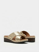 Gold textured cross-strap wedge mule sandals with espadrille trim and dark wood-look wedge sole, close angled view.