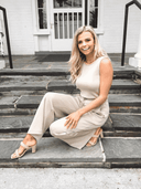 Blonde woman in a beige sleeveless jumpsuit, crouching on stone steps, angled front view.