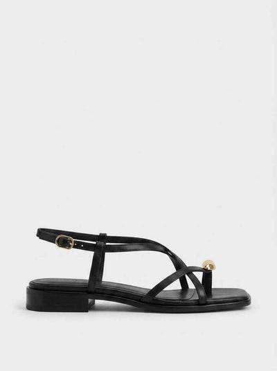 Black strappy flat sandal with crossover toe straps, gold orb accent and ankle buckle, side profile on white background.