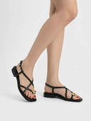Black strappy flat sandals with gold orb detail worn on feet, showing crossover toe straps and ankle buckle, angled view.