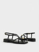 Black strappy flat sandals showing gold buckle ankle strap and gold orb toe detail, angled view on white background.