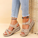 Beige low wedge platform sandals with adjustable velcro strap bands and contrast stitching, worn with distressed jeans — angled front view.