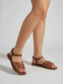 Brown flat crisscross-strap sandals with elastic ankle band worn on feet, angled lifestyle view.
