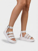 White chunky flatform sandals showing silver square buckles and rows of metal eyelets on multi-strap upper, angled three-quarter view.