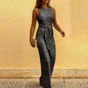 Woman in black sequin jumpsuit with tie waist, holding a clutch, angled view on yellow background.