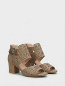 Taupe laser-cut block heel sandal showing ankle buckle fastening and geometric cut-out panel, angled side view on white background.
