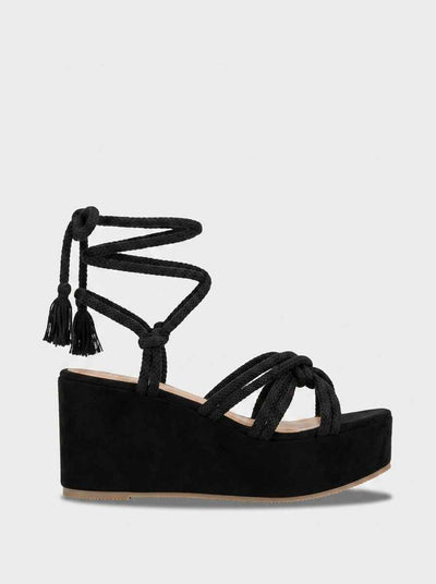 Black platform wedge sandals with braided rope lace-up straps and tassel ankle ties, side view on white background.