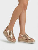 Rose gold metallic espadrille wedge sandals with jute rope platform sole and stud trim, worn on feet, angled view.