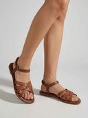 Brown flat crisscross strap sandals with elastic ankle strap worn on feet, angled view.