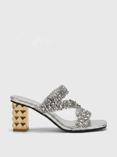 Silver sequin crossover strap block heel mules with gold pyramid-studded heel, side profile view on white background.