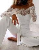 White off-shoulder lace-accent jumpsuit with long sleeves, worn by a person sitting on a wooden floor.